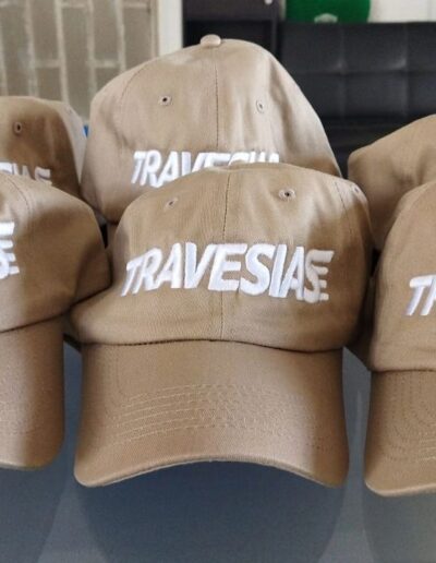 Grupo de gorras color beige con la palabra 'TRAVESIAS' bordada en blanco.